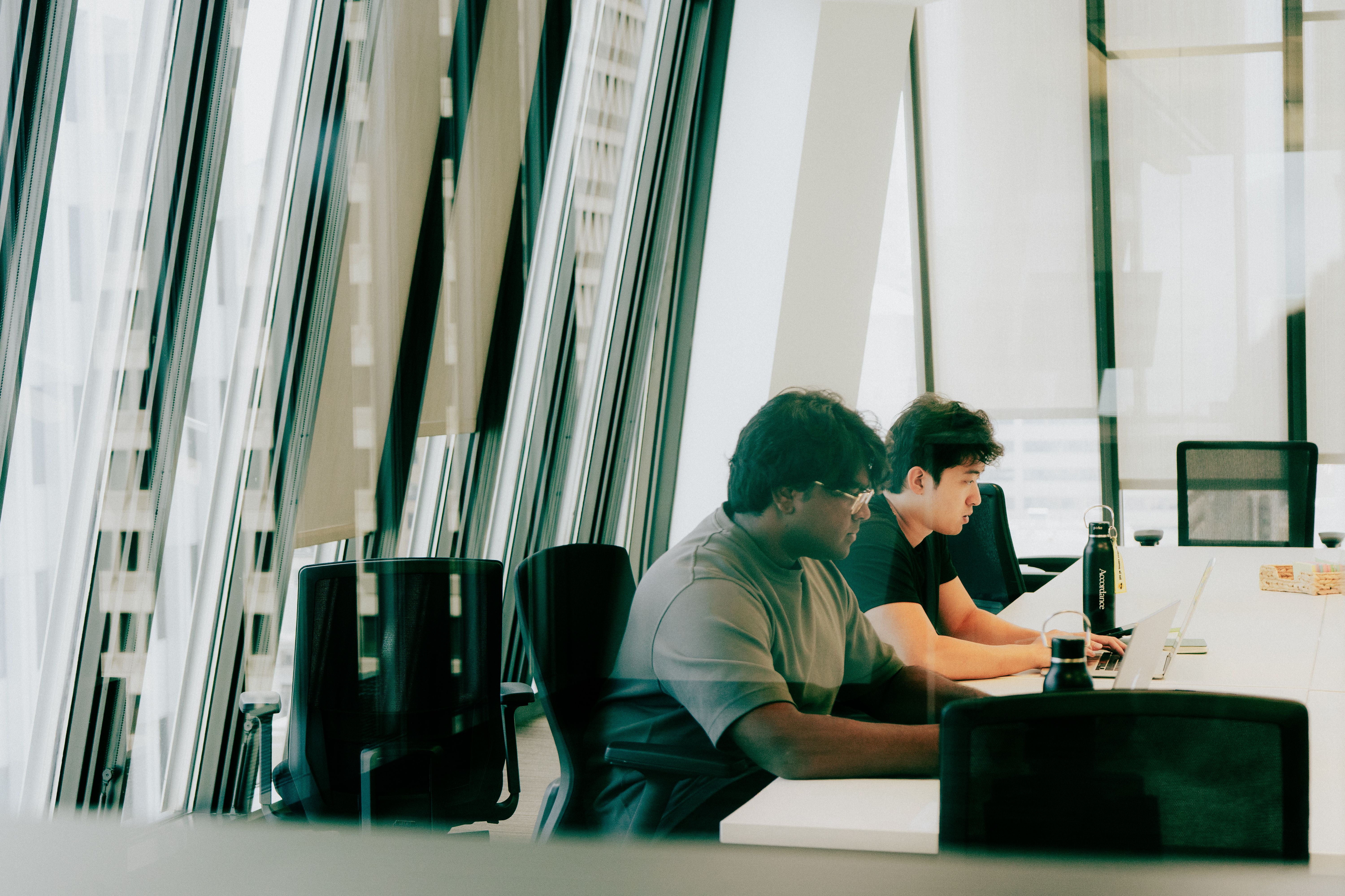 Two men work on laptops at a white table in a modern office with large windows.