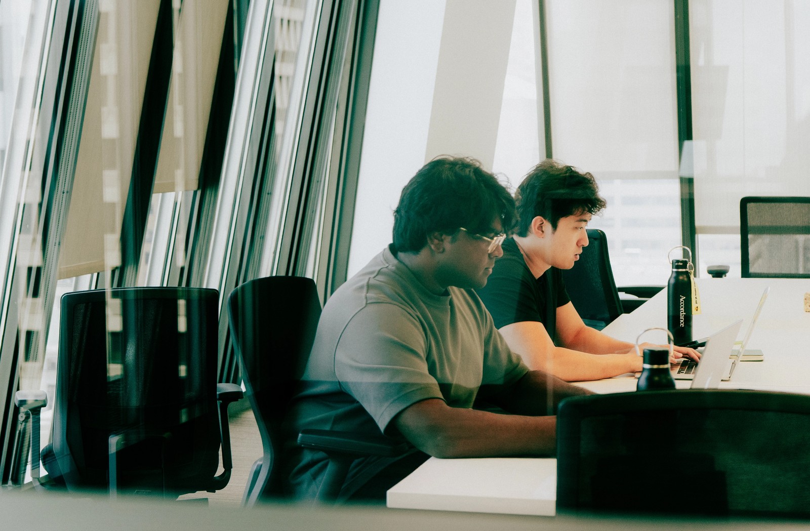 Two men work on laptops at a white table in a modern office with large windows.