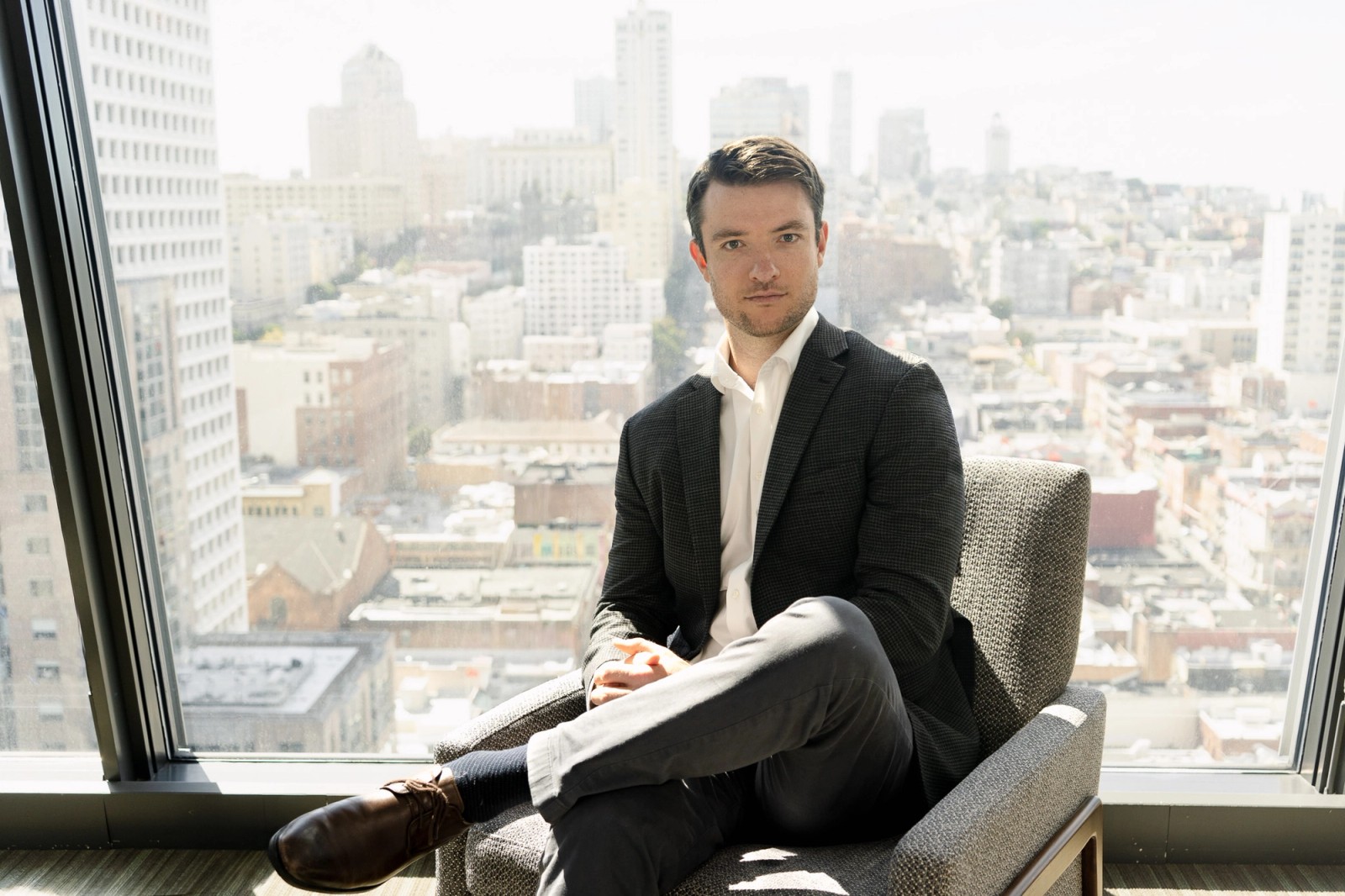 A man in a suit sits in a chair by a large window overlooking a city skyline.