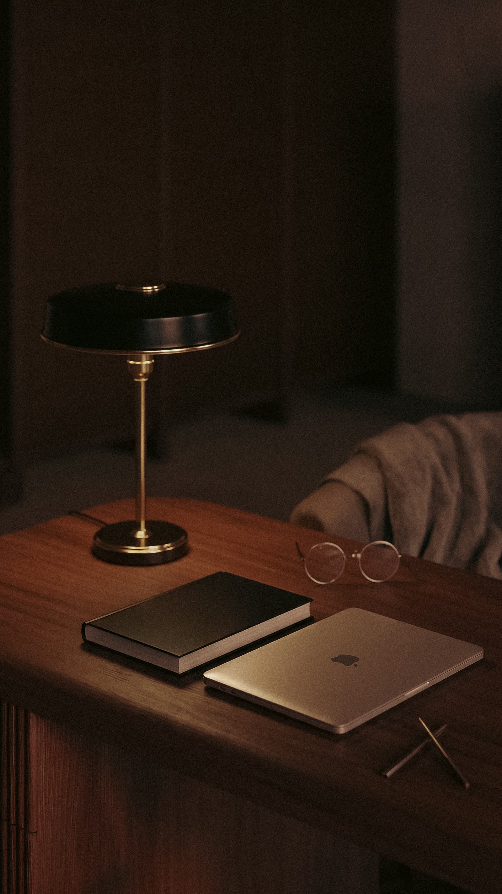 A dimly lit wooden desk with a vintage black and gold lamp, a closed silver laptop, a black book, and reading glasses.