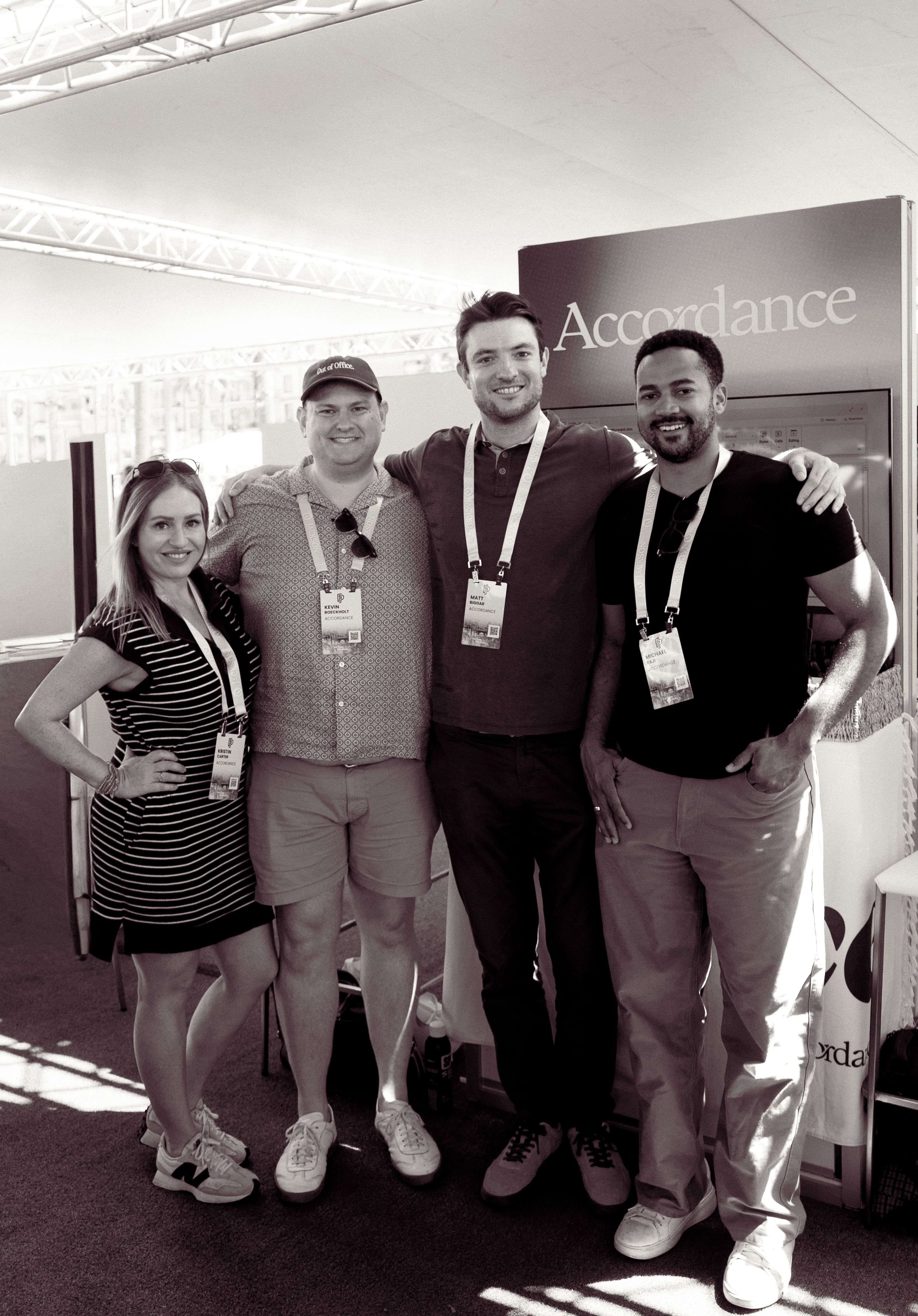 Four smiling people, one woman and three men, wearing lanyards at an "Accordance" event booth.