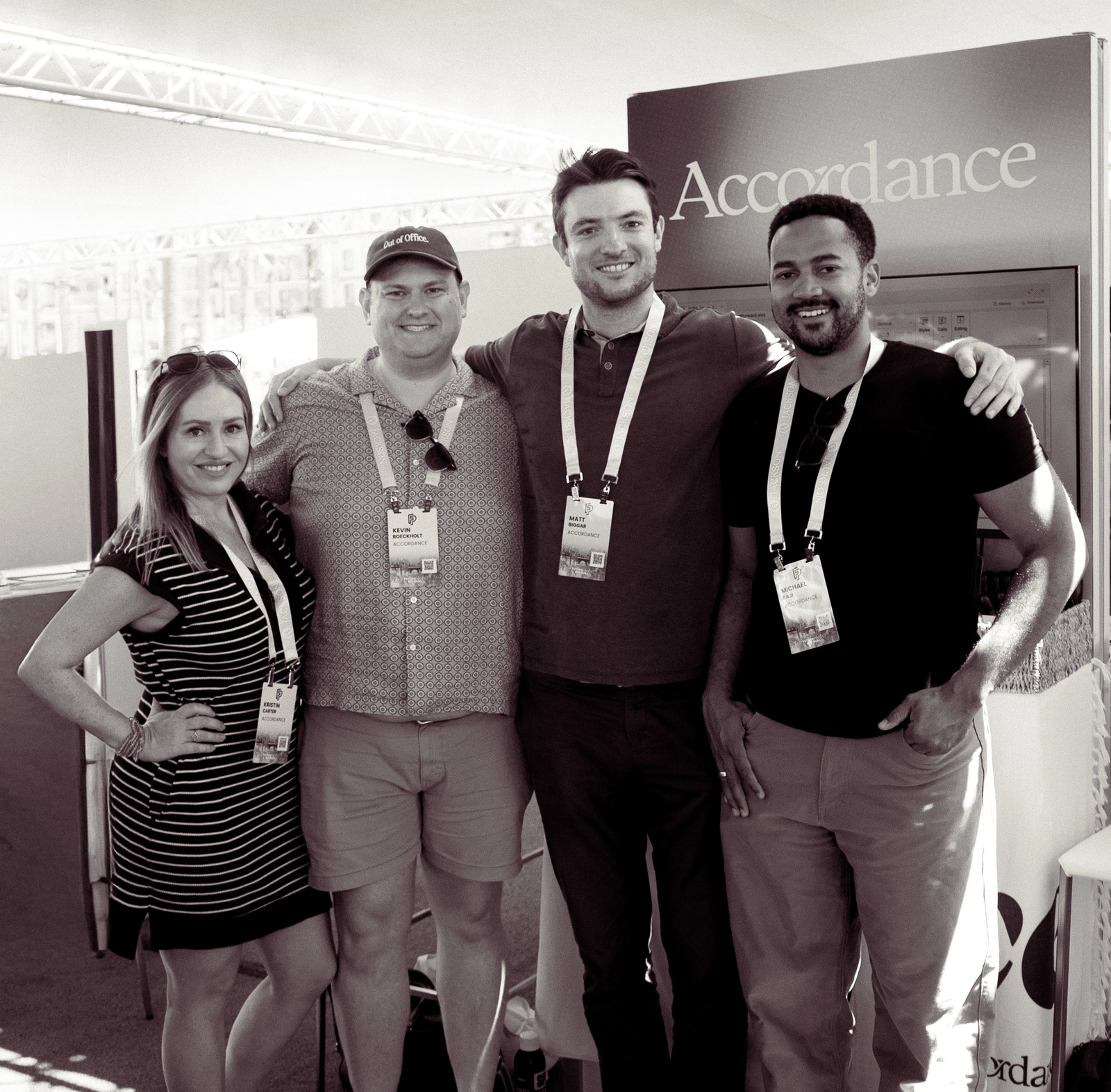 Four smiling people, one woman and three men, wearing lanyards at an "Accordance" event booth.