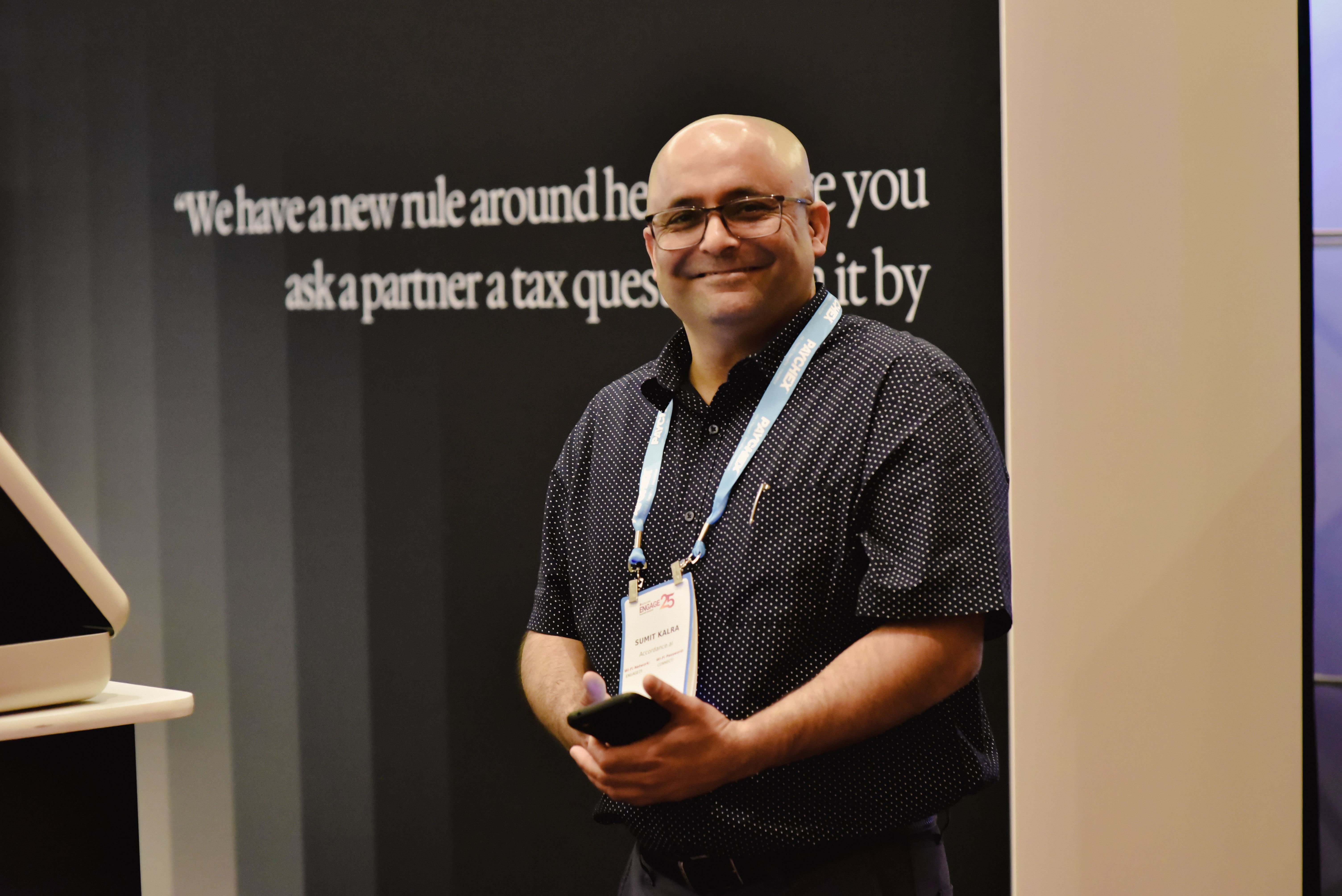 Smiling man in glasses and an event lanyard holds a phone in front of a wall with text.