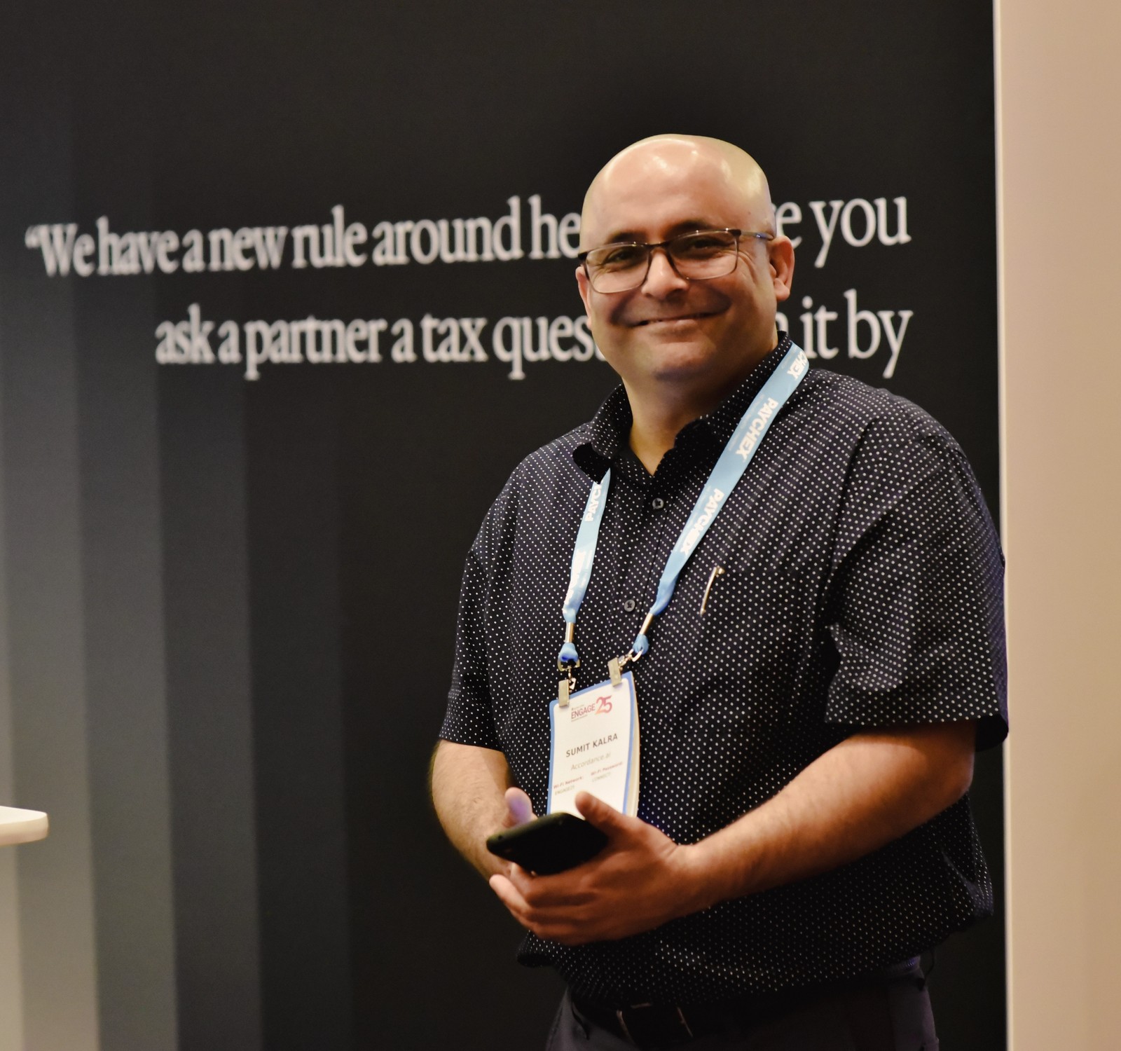 Smiling man in glasses and an event lanyard holds a phone in front of a wall with text.