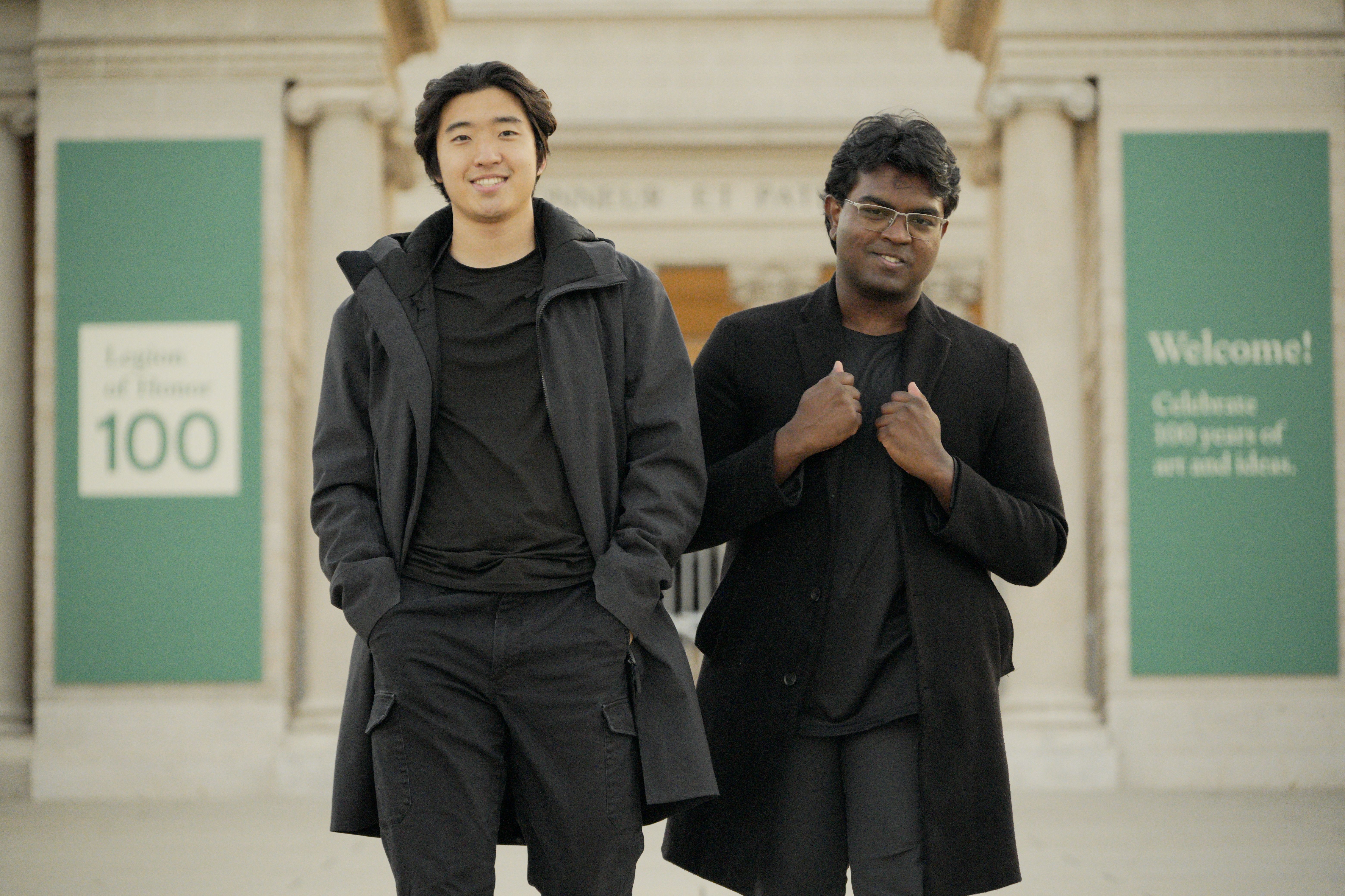 Two smiling men, one East Asian and one South Asian, wearing dark coats, stand in front of a building with columns and green banners.