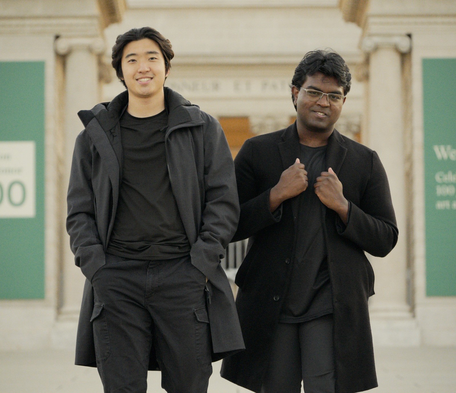 Two smiling men, one East Asian and one South Asian, wearing dark coats, stand in front of a building with columns and green banners.