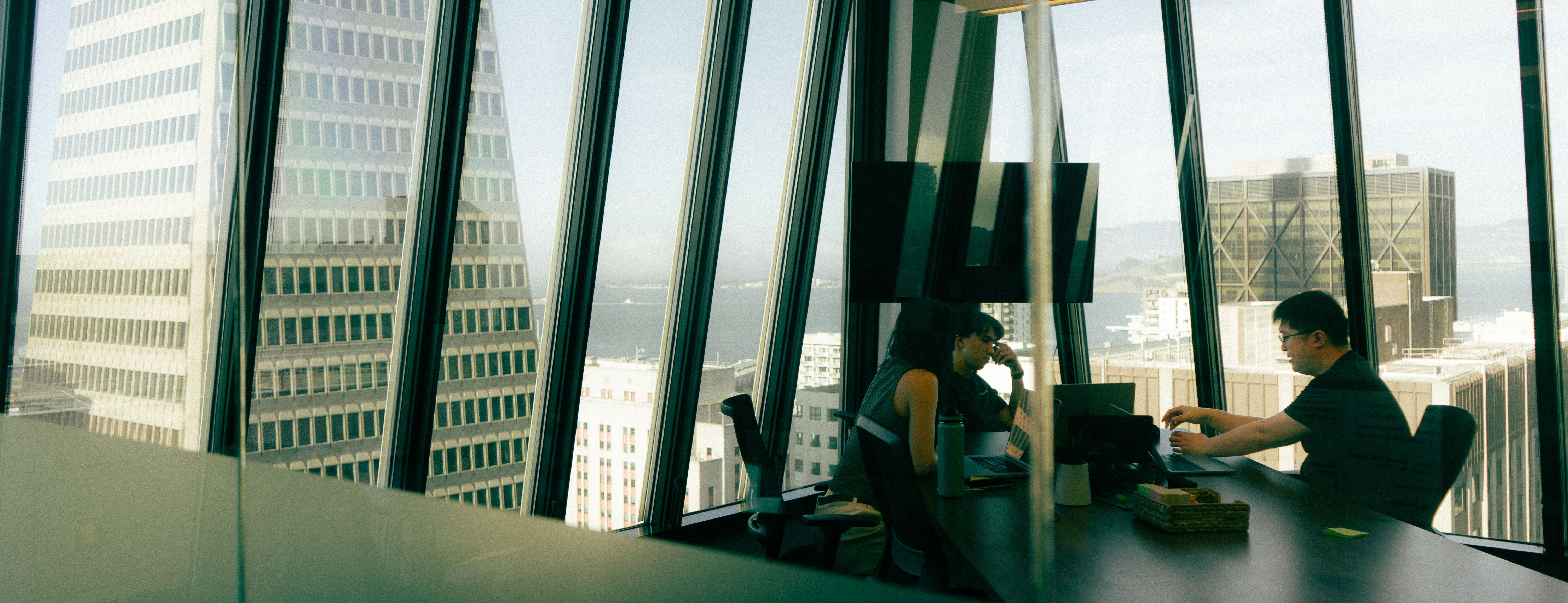Three people working at a desk in a modern high-rise office with large, angled windows overlooking a city and bay.