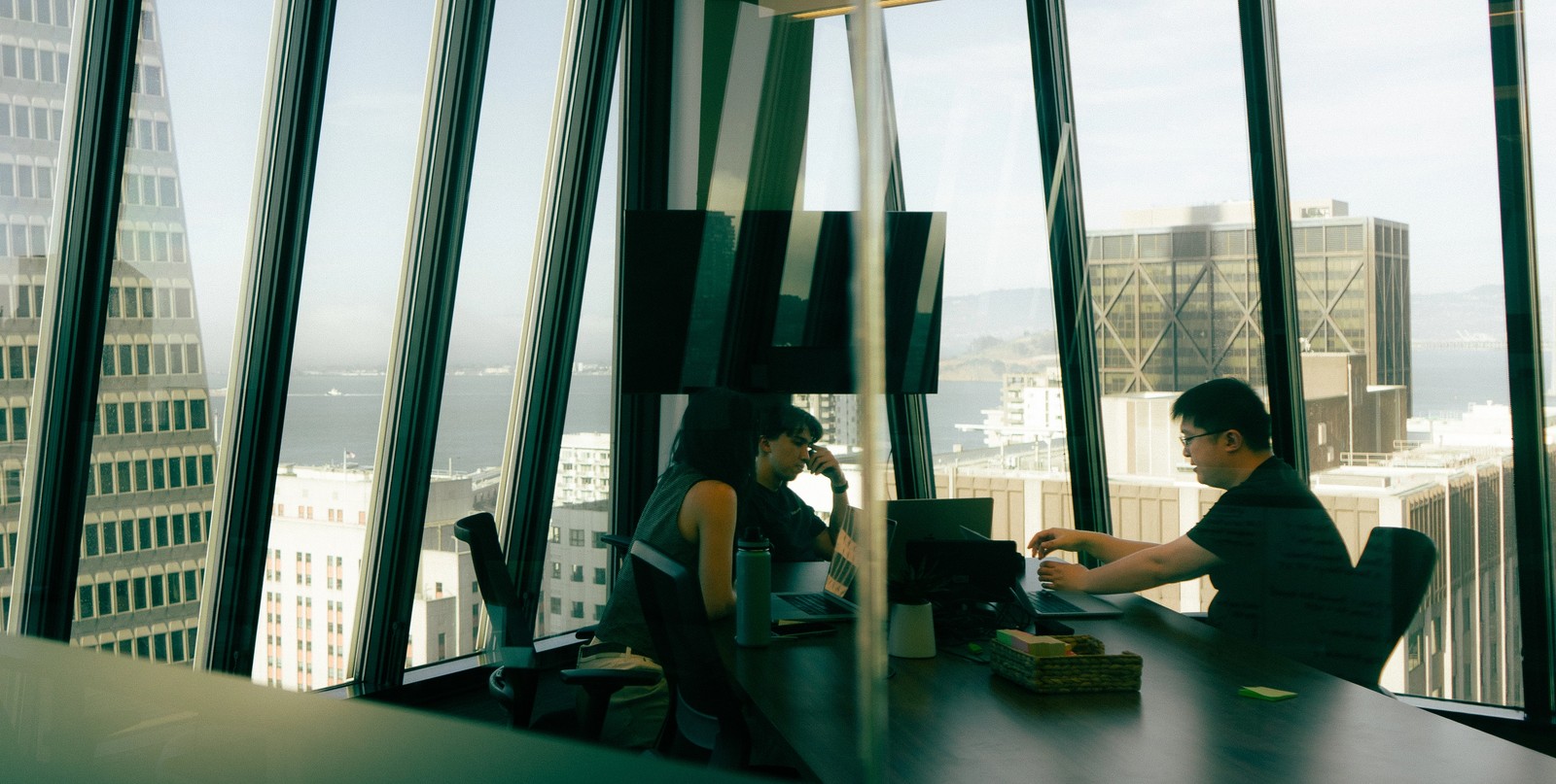 Three people working at a desk in a modern high-rise office with large, angled windows overlooking a city and bay.