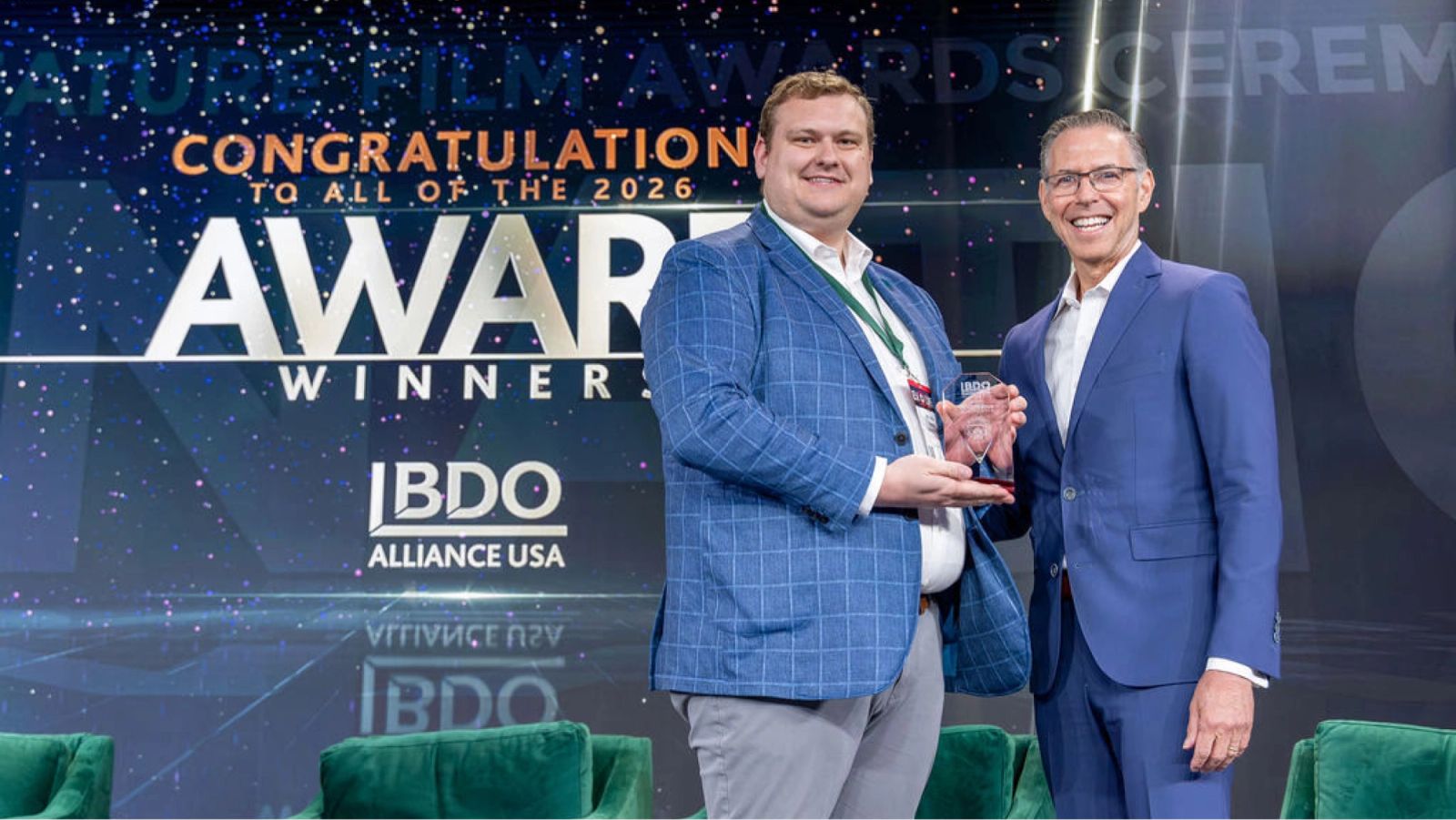 Two smiling men on stage; one in a blue plaid jacket accepts a clear award from another in a blue suit, with "AWARD WINNER" and "BDO ALLIANCE USA" on the backdrop.