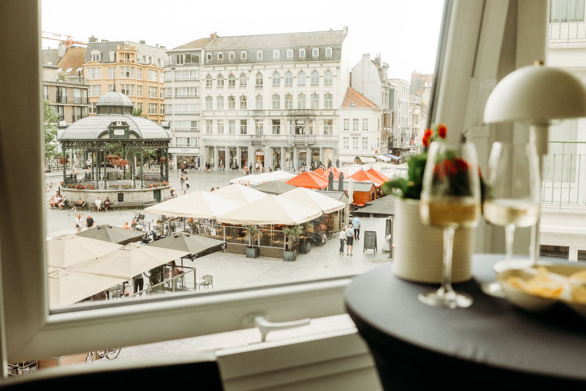 Zicht vanuit het raam op het Wapenplein met de kiosk en de terrassen, met twee glazen wijn op de statafel