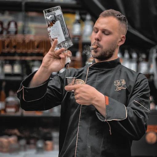 A stylish bartender in a black uniform pouring a drink into a shot glass while puffing on a cigar.