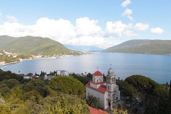 The historic Savina Monastery, an Orthodox church with a red dome, nestled in greenery overlooking the scenic Bay of Kotor in Herceg Novi