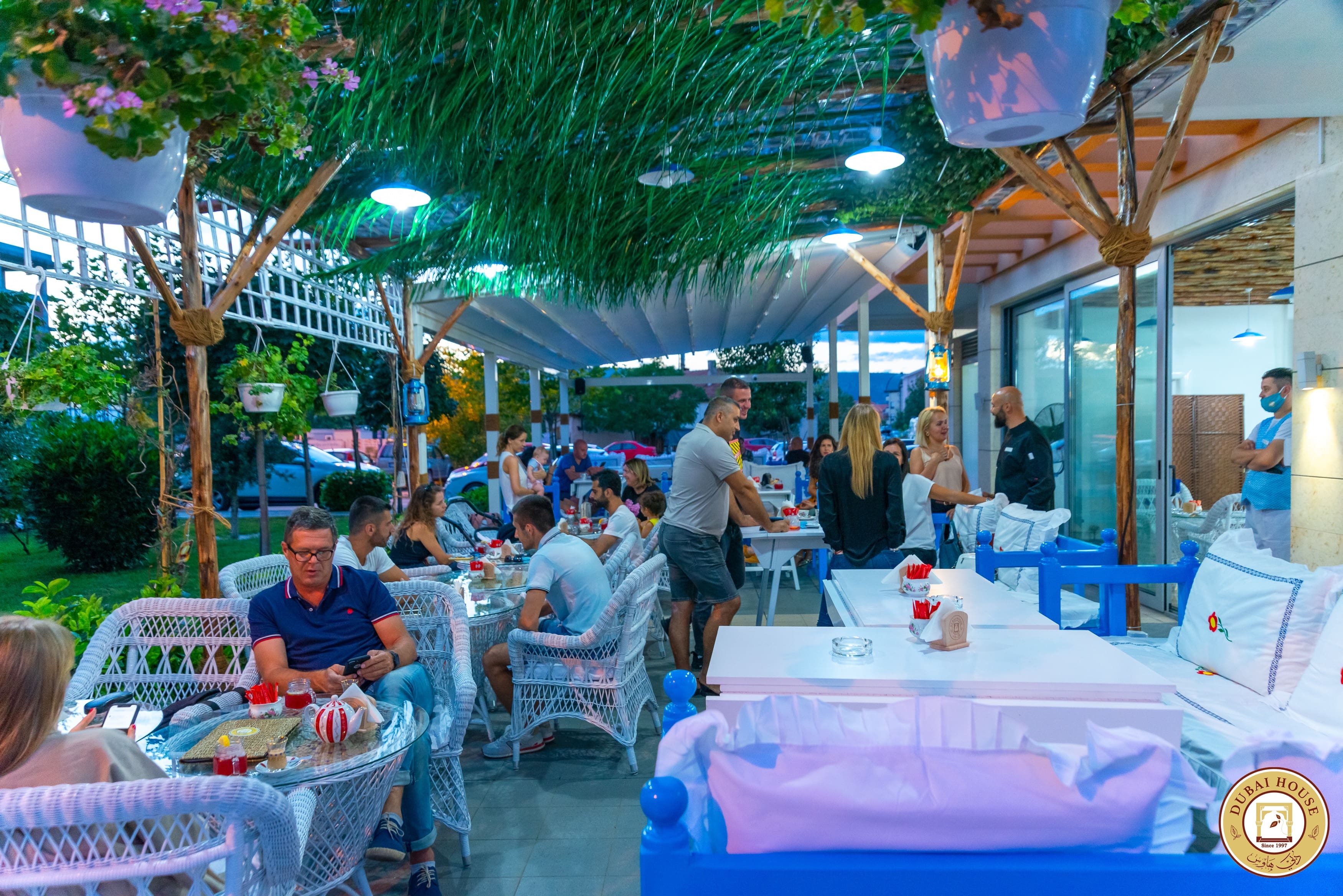 The vibrant and bustling outdoor terrace of the Arabian Tea House in Podgorica, filled with guests dining at white tables.
