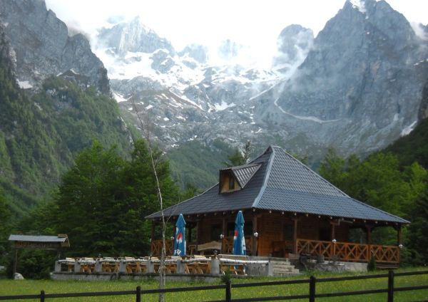 A rustic wooden mountain lodge in the Grebaje Valley, Montenegro, dwarfed by the dramatic, jagged, and snow-dusted peaks of the Prokletije mountains