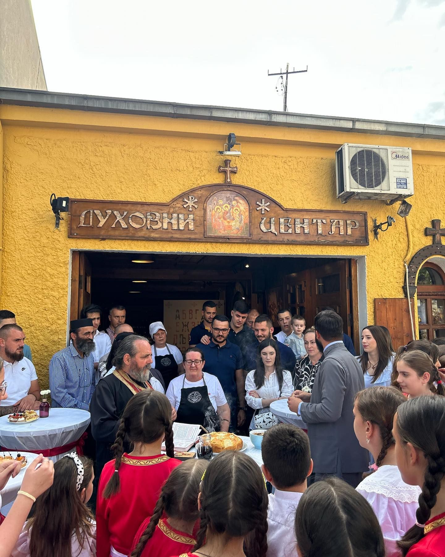 An Orthodox priest conducting a blessing ceremony with a loaf of bread in front of the Duhovni Centar building, surrounded by a crowd.
