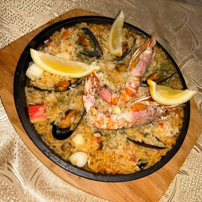 A sizzling black pan filled with seafood paella, featuring prawns, mussels, and calamari, garnished with lemon wedges.