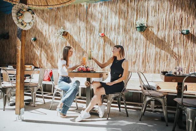 Two women toasting with cocktails at a table in a sunny, rustic-chic patio with a woven bamboo wall.