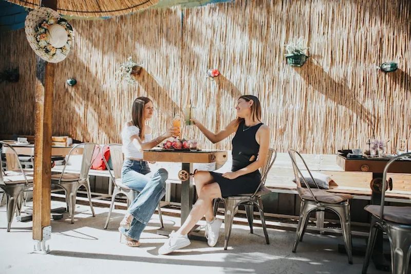 Two women toasting with cocktails at a table in a sunny, rustic-chic patio with a woven bamboo wall.