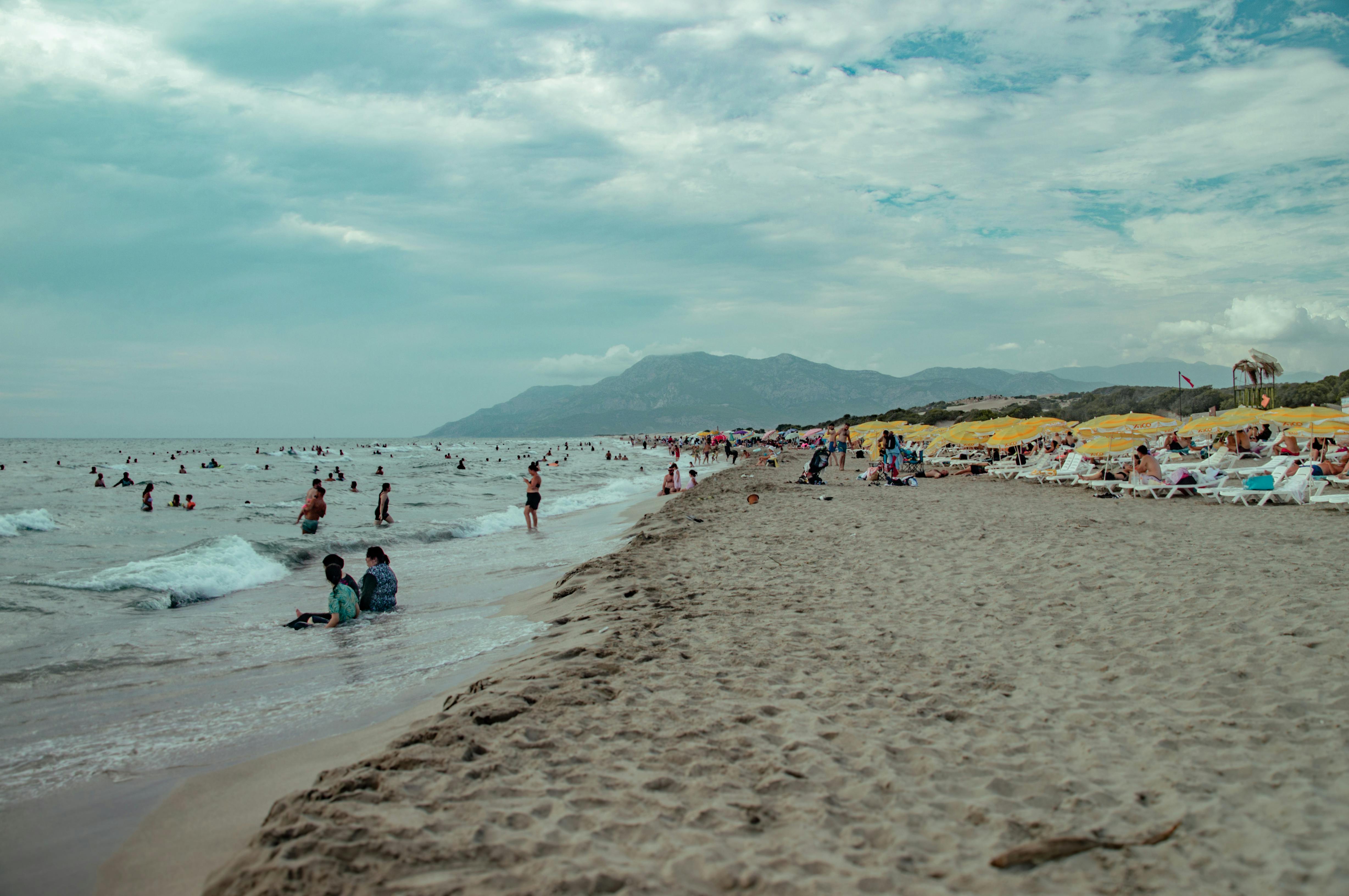 People swimming and relaxing on the wide sandy shore of Velika Plaža on a cloudy day, with mountains in the background.