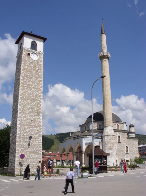 The historic Husein Pasha's Mosque in Pljevlja, Montenegro, with its tall minaret and domes, standing next to a stone clock tower on a sunny day