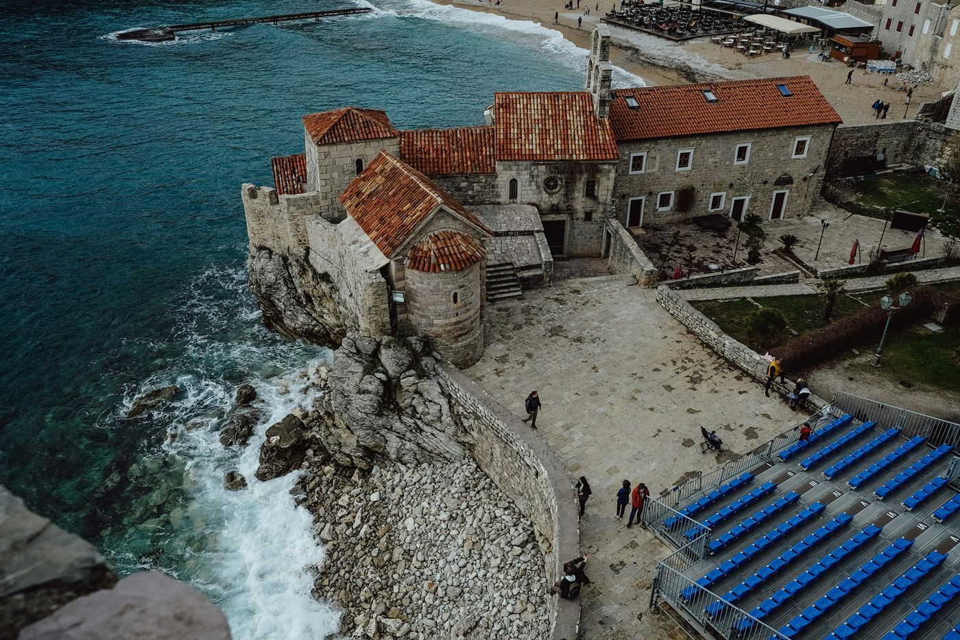 A high-angle view of the Budva Citadel in Montenegro, showing the historic stone Church of Santa Maria in Punta on a rocky cliff overlooking the sea