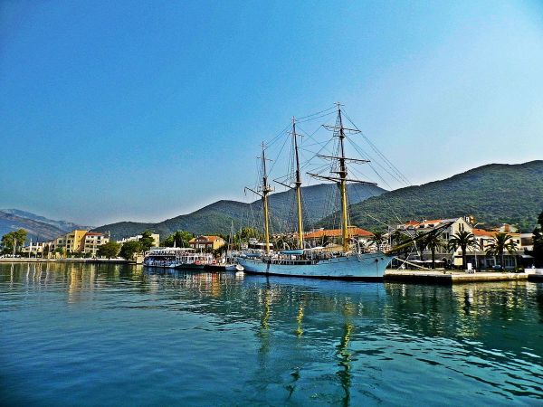 Ein großes, weißes, dreimastiges Großsegelschiff, das Schulschiff Jadran, das am Ufer von Tivat angedockt ist, mit Palmen, Gebäuden und grünen Bergen im Hintergrund