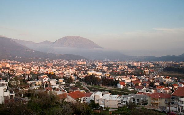 A panoramic view of the modern city of Bar, Montenegro, bathed in the warm light of sunrise, with large mountains shrouded in mist in the background