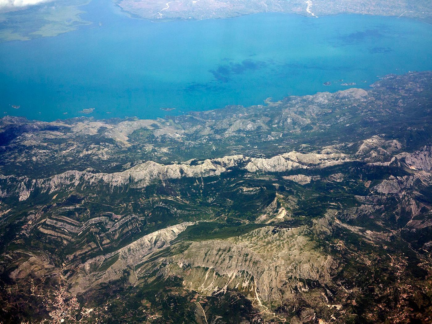 An aerial photograph of the vast blue waters of Skadar Lake, separated from the coast by dramatic, rugged mountains.