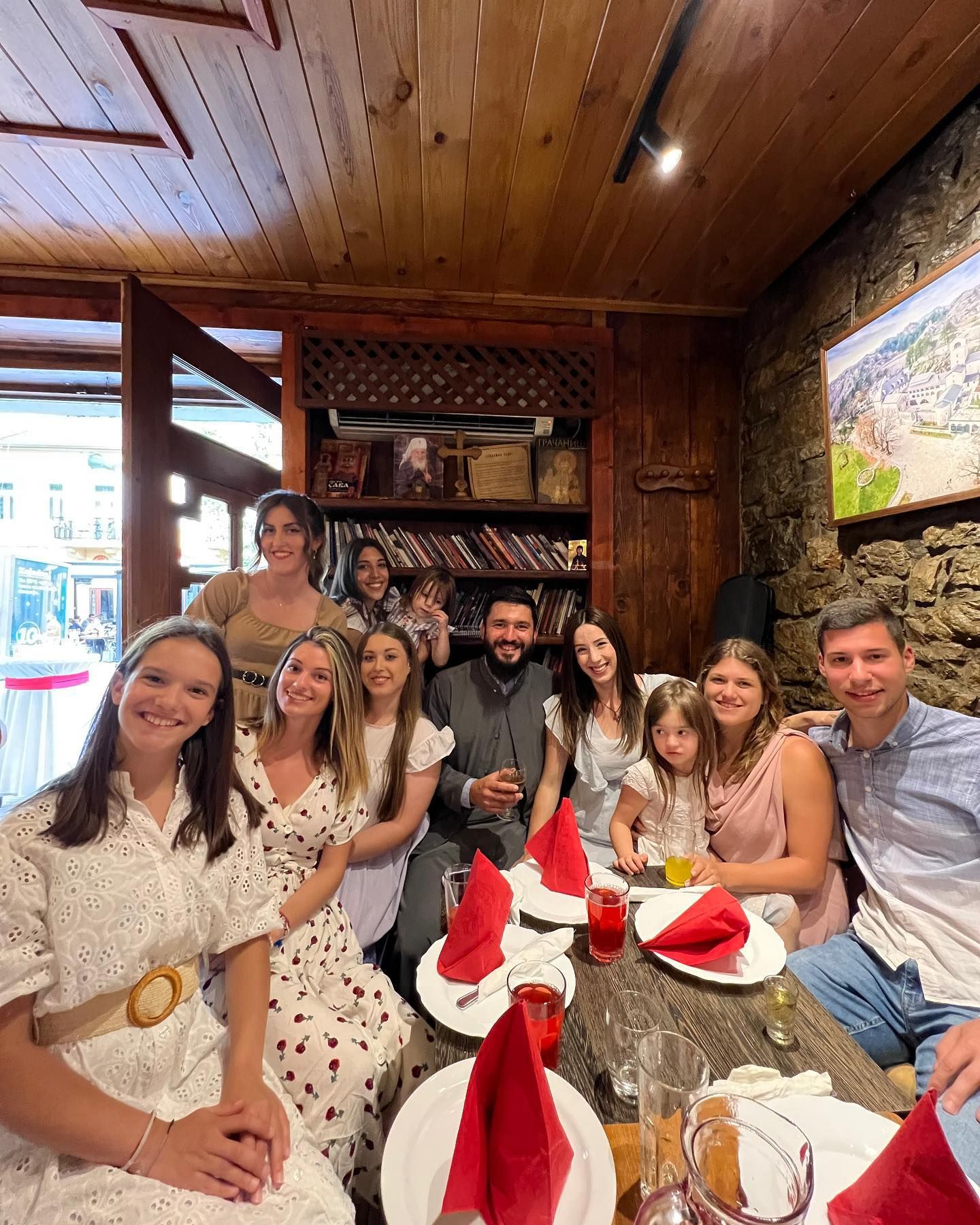 A large, happy group of people, including a priest, gathered around a long wooden table for a celebratory meal inside a rustic restaurant.