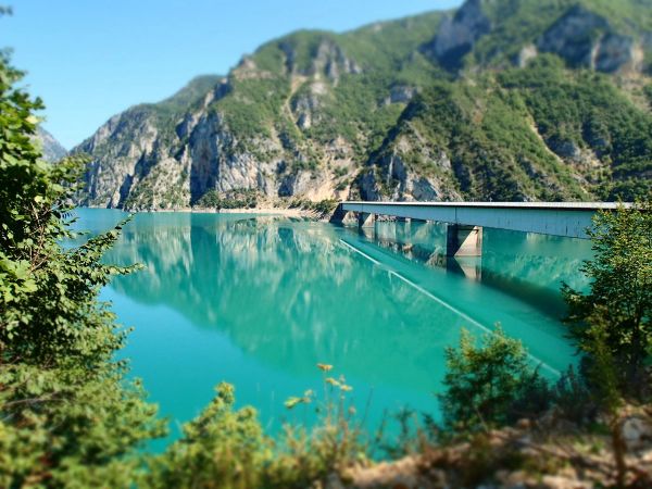 A long bridge crossing the calm, turquoise water of Lake Piva in Montenegro, with the surrounding green mountains perfectly reflected on the lake's surface