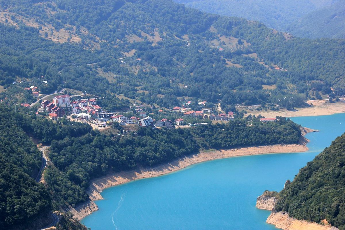 A scenic, high-angle view of the town of Plužine, nestled on the shore of the stunningly turquoise Lake Piva, surrounded by green mountains in Montenegro