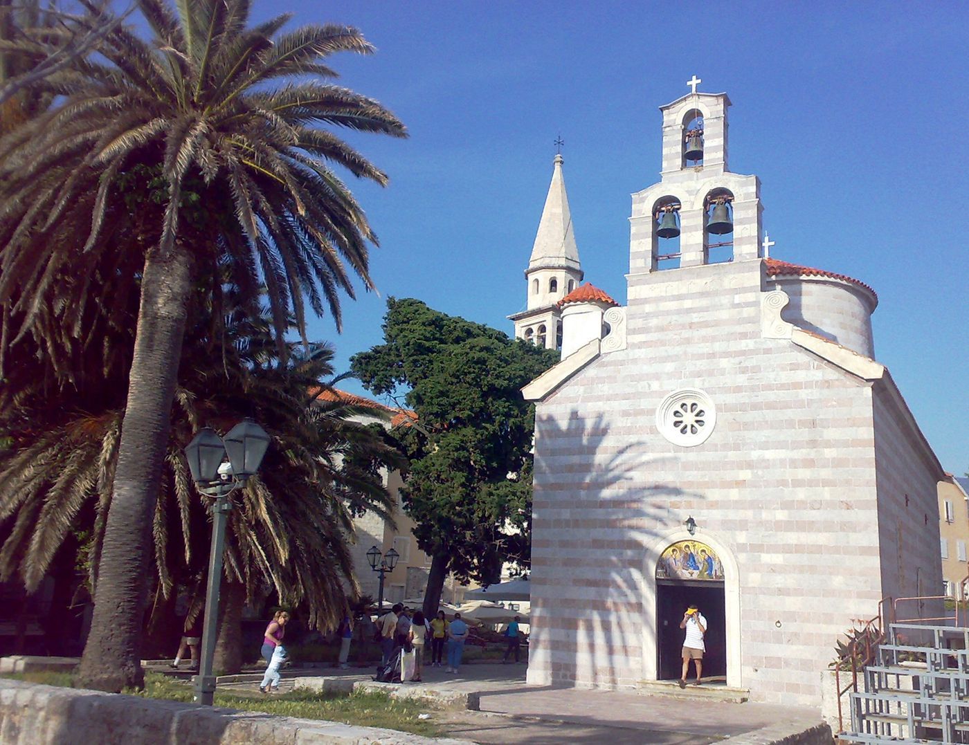 Церковь Святой Троицы в Старом городе Будвы с колокольней и пальмой на переднем плане.