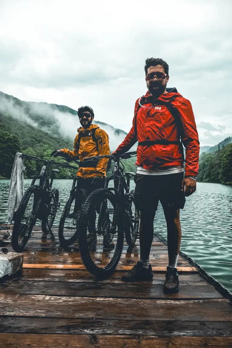 Two mountain bikers in rain jackets standing with their bikes on a wooden dock by a misty lake, surrounded by mountains.