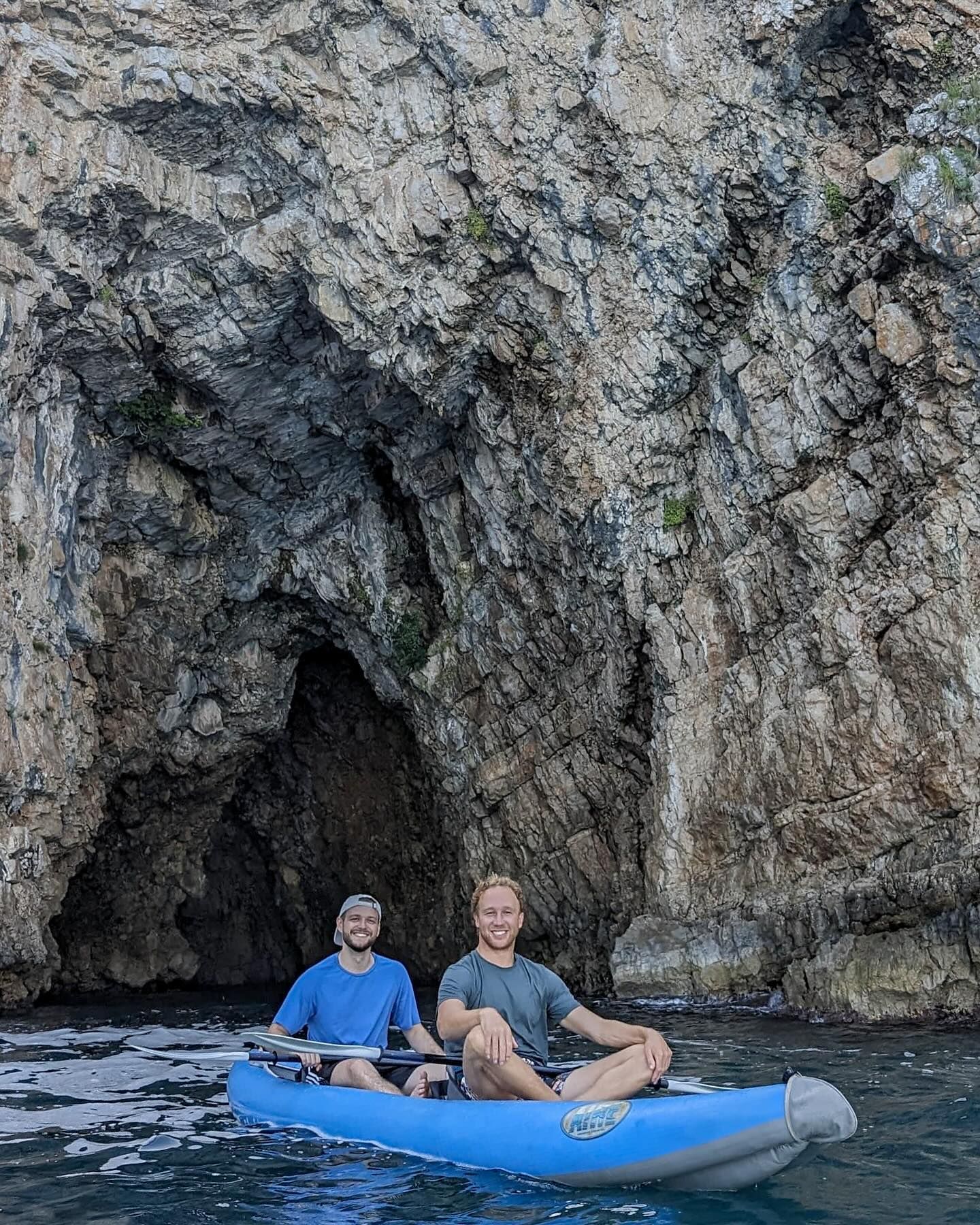 Dva muškarca se smiju u plavom gumenom kajaku na ulazu u veliku, stjenovitu morsku pećinu.