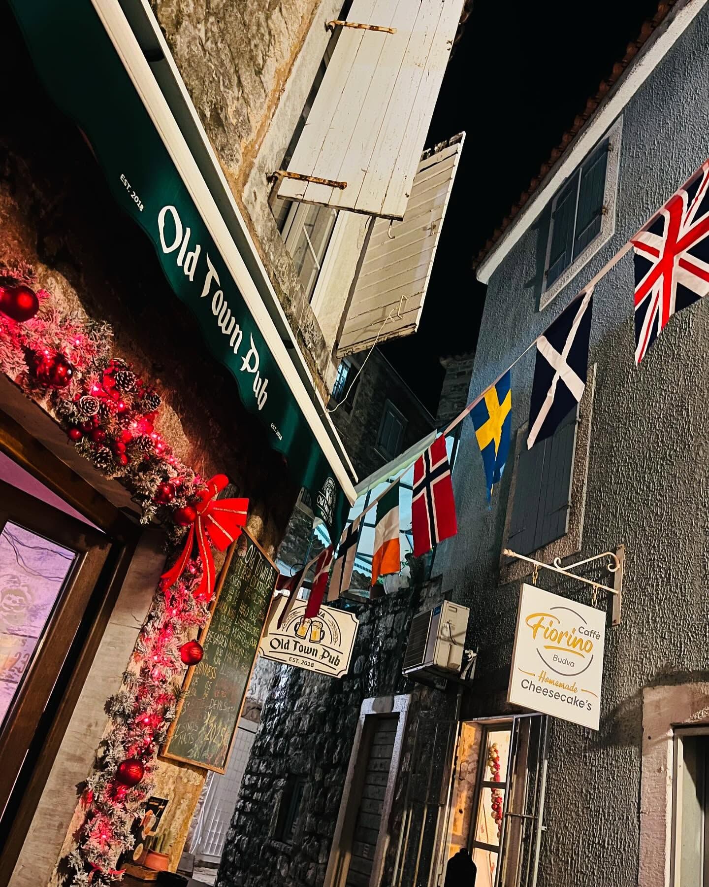 A narrow stone alley in Budva's Old Town at night, with the "Old Town Pub" sign illuminated and various national flags hanging above.