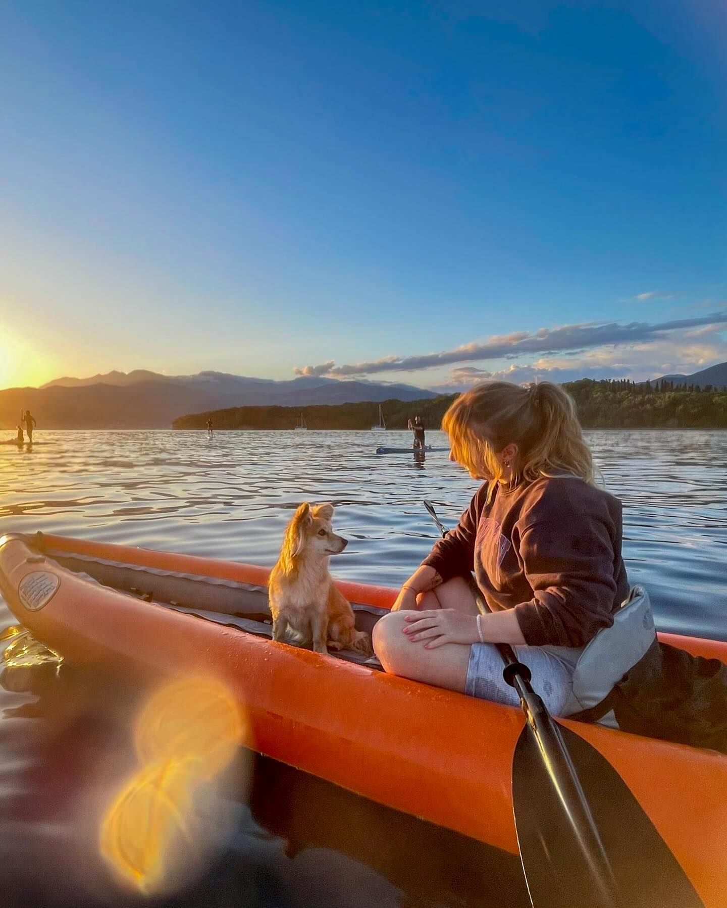 Žena i njen mali pas sjede u narandžastom kajaku na vodi u zalazak sunca, sa planinama u pozadini.