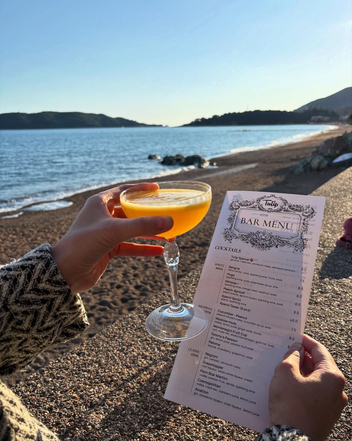 A person holding a vibrant orange cocktail on a beach, with the Restaurant TULIP bar menu and the sea in the background.