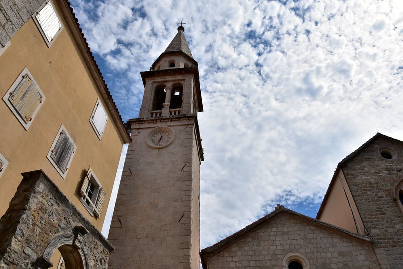 A low-angle view looking up at the stone bell tower of St. John's Church in Budva against a cloudy sky.