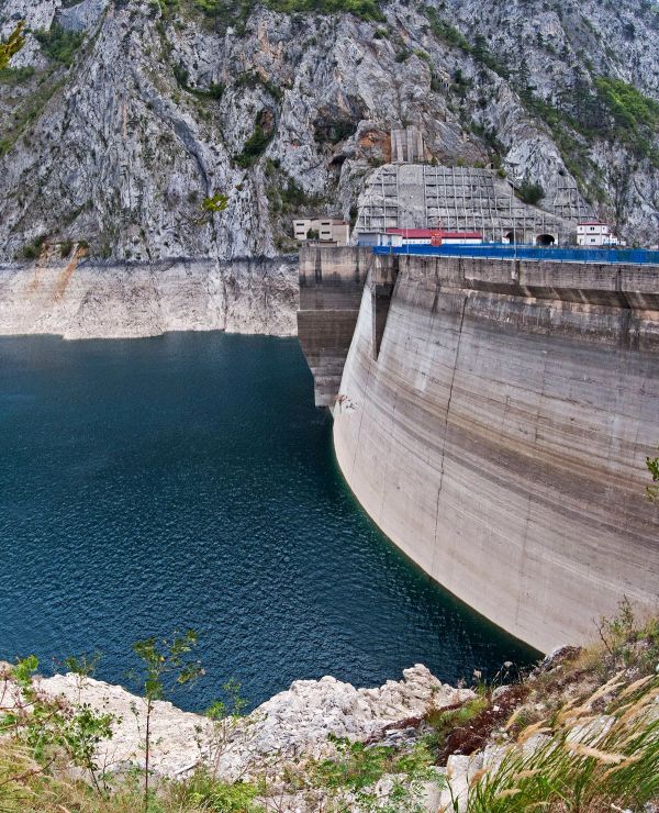 A close-up view of the immense, arched concrete wall of the Mratinje Dam in Montenegro, holding back the deep blue water of Lake Piva within a steep rock canyon