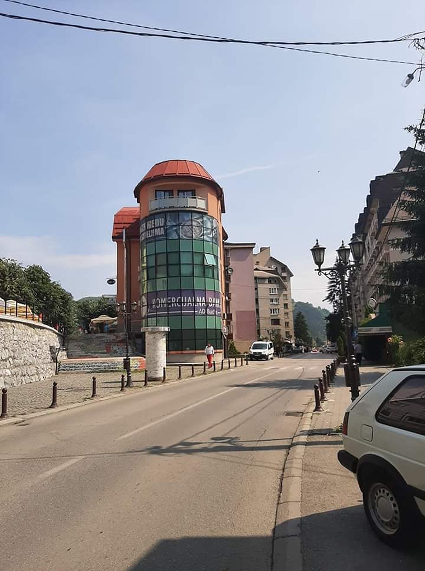 A street scene in Bijelo Polje, Montenegro, focusing on a modern building with a curved glass facade and a red-roofed circular tower