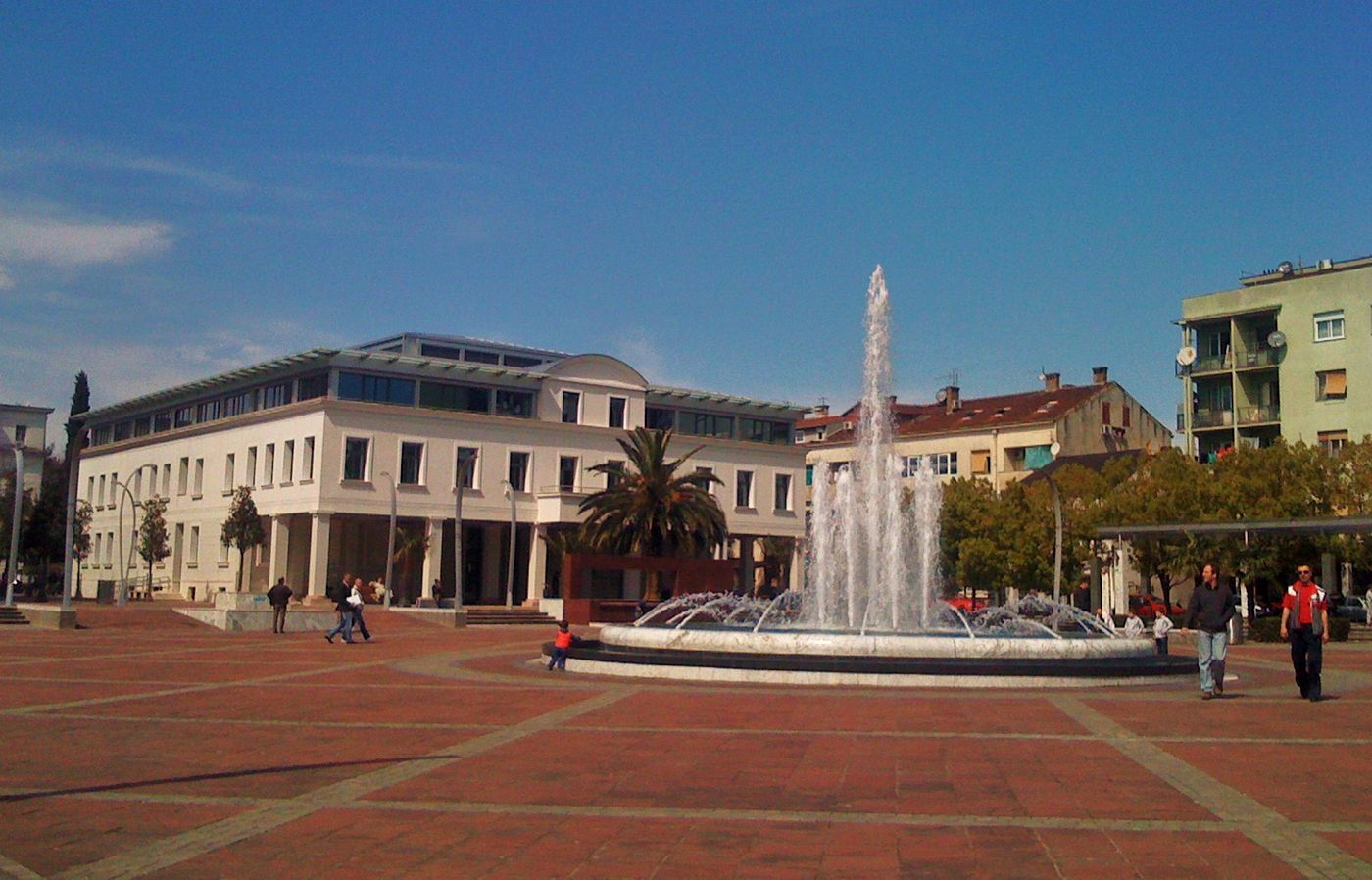 People walking around the central fountain of Independence Square in Podgorica under a clear blue sky.