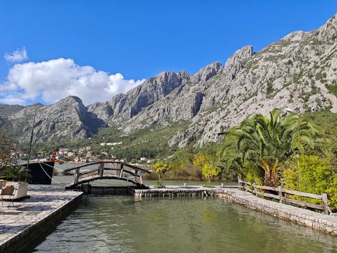 Scenic view from Stari Mlini showing its waterfront dock, a quaint wooden bridge, and the majestic mountains rising above the Bay of Kotor under a clear blue sky.