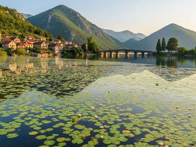 Serene view of Lake Skadar covered in water lilies, with the village of Virpazar and a stone bridge in the background, surrounded by mountains.