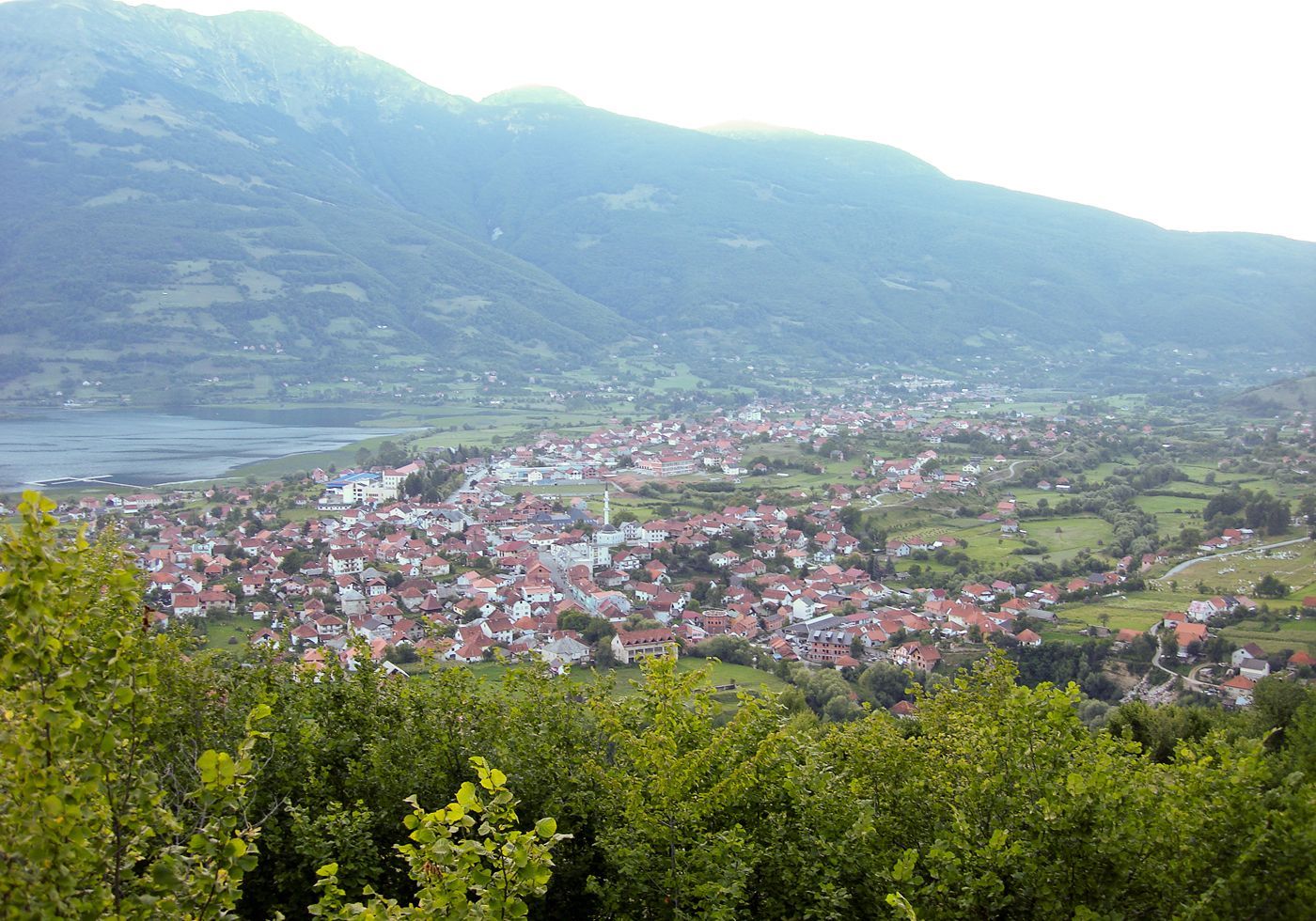 A panoramic view from a hill overlooking the town of Plav, Montenegro, showing the red-roofed houses, the adjacent Lake Plav, and the vast mountain valley of Prokletije