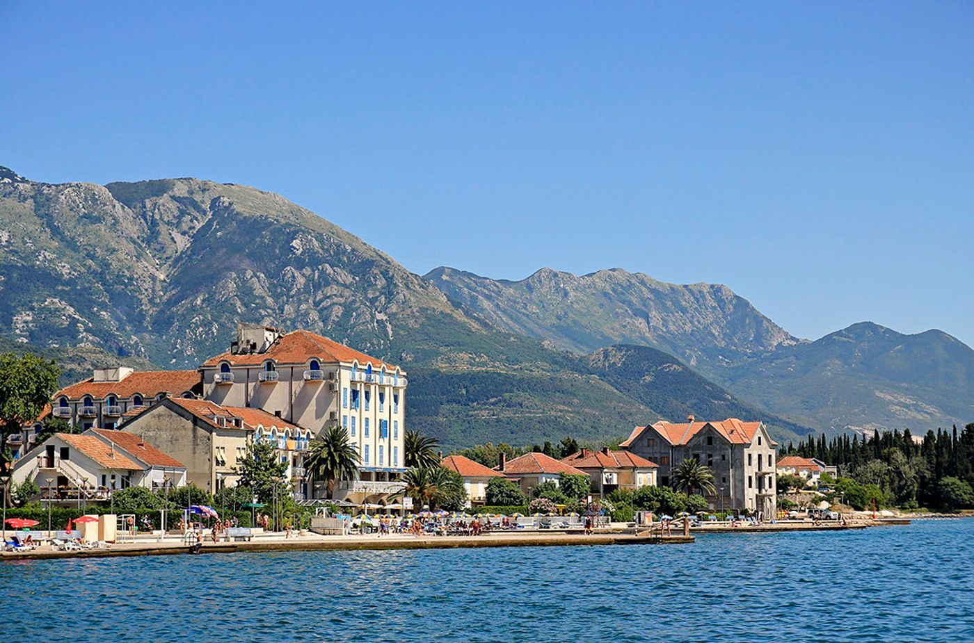 A view from the water of the Tivat waterfront in Montenegro, showing a historic hotel, stone houses, and a beach promenade at the foot of large mountains