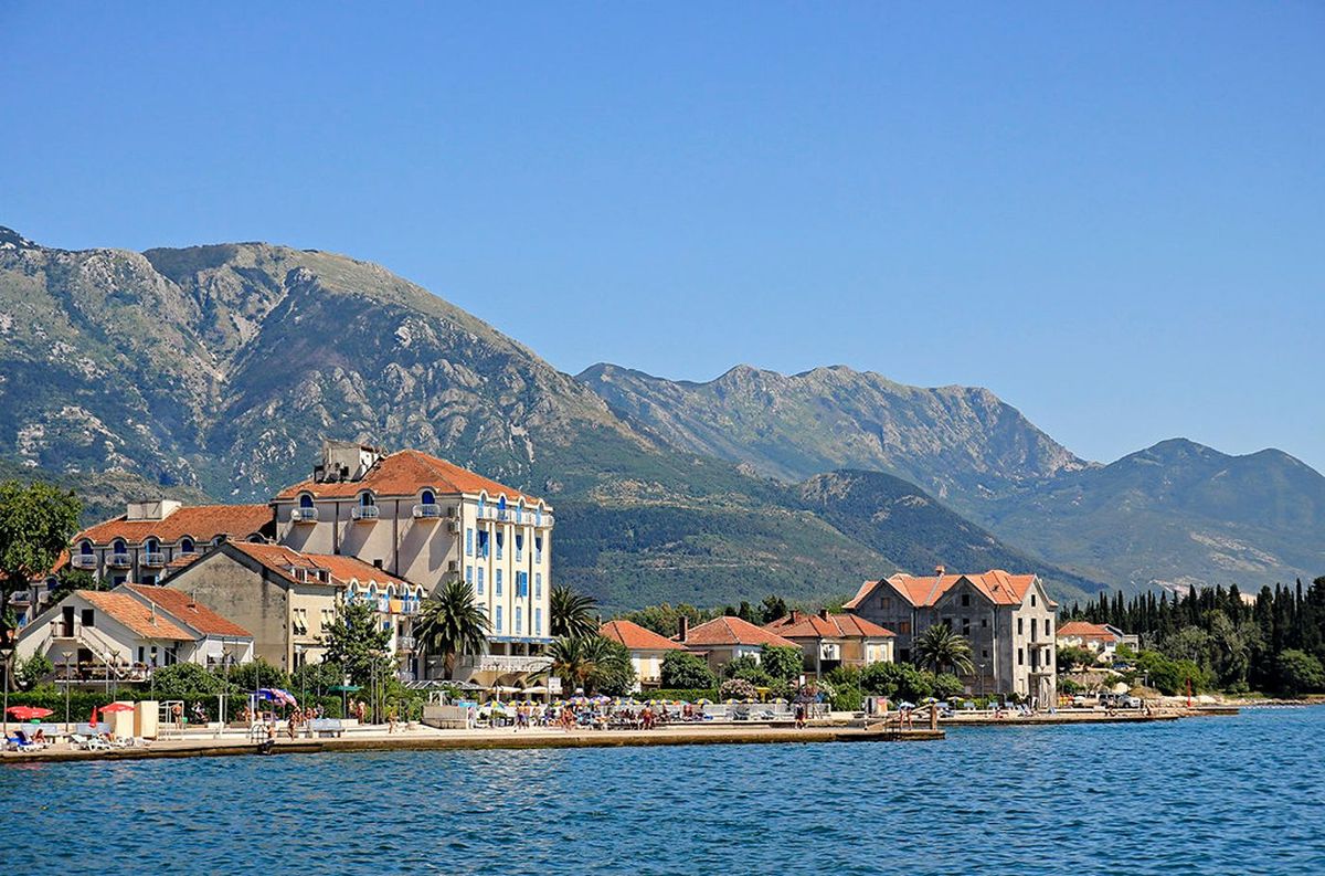 Ein Blick vom Wasser auf die Uferpromenade von Tivat, Montenegro, mit einem historischen Hotel, Steinhäusern und einer Strandpromenade am Fuße großer Berge