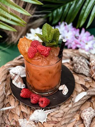 A vibrant, fruity cocktail garnished with raspberries and mint, presented on a woven mat with seashells.