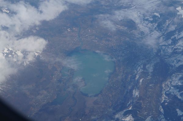 A high-altitude aerial view of Lake Plav in Montenegro during winter, showing the turquoise lake surrounded by valleys, with snow-covered mountains on the right and scattered clouds