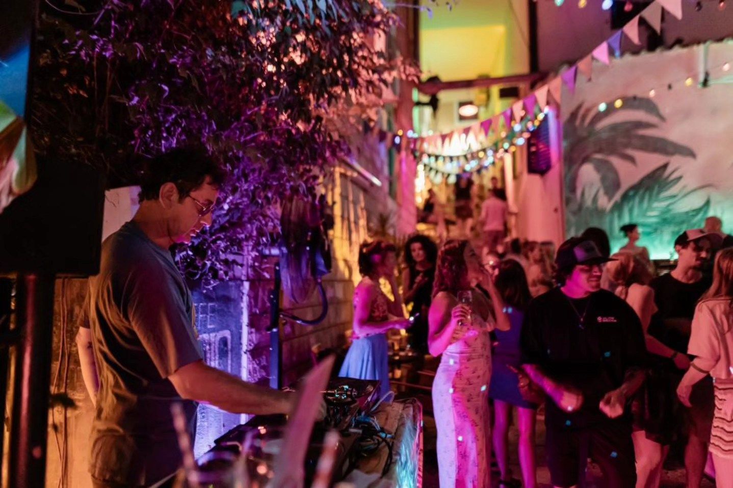 A DJ playing music for a lively crowd dancing in a colorful, plant-filled outdoor patio at night.