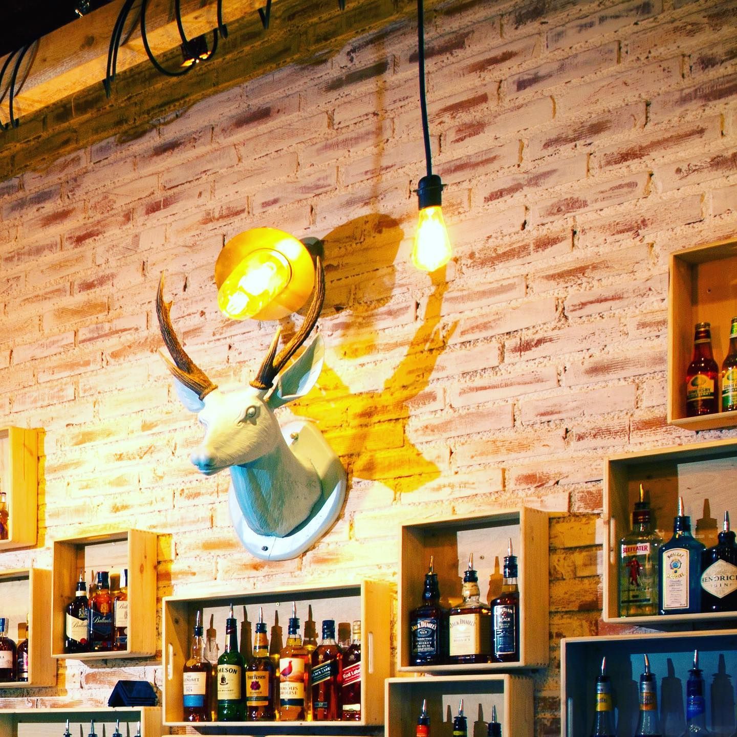A decorative white deer head mounted on a brick wall, surrounded by shelves of premium spirits in wooden boxes, creating a rustic-chic vibe.